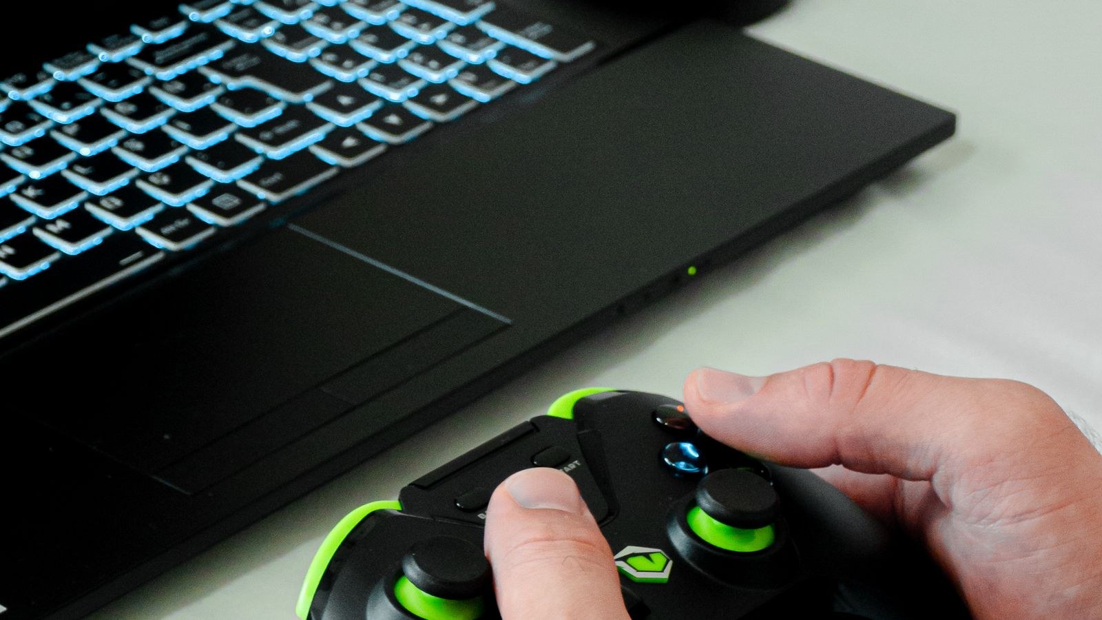 Hands holding a wireless gaming controller in front of a laptop with a backlit keyboard