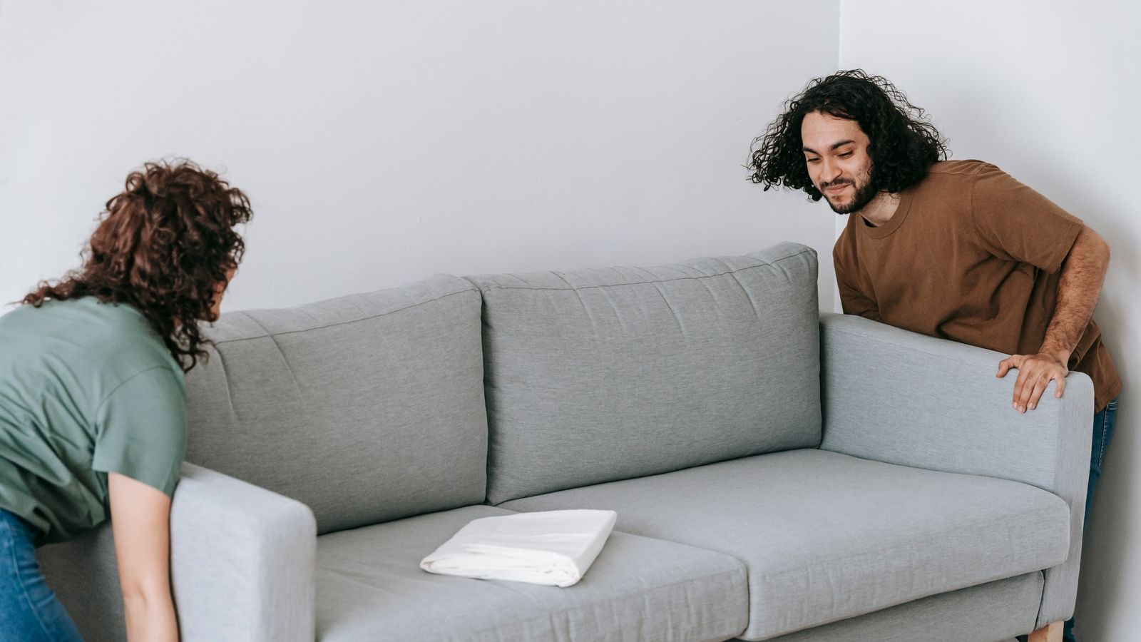 Two people repositioning a gray couch in a living room during furniture rearrangement