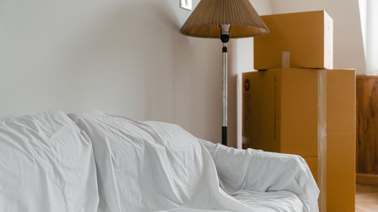 Sofa covered with a protective sheet beside stacked cardboard moving boxes in a bright room