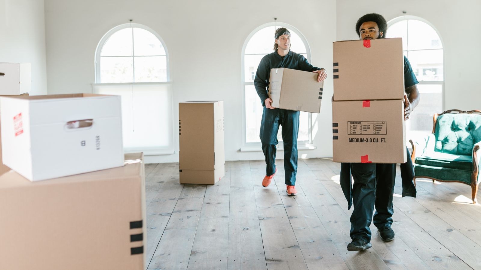 Two movers carrying wrapped boxes and furniture through a sunlit room on moving day