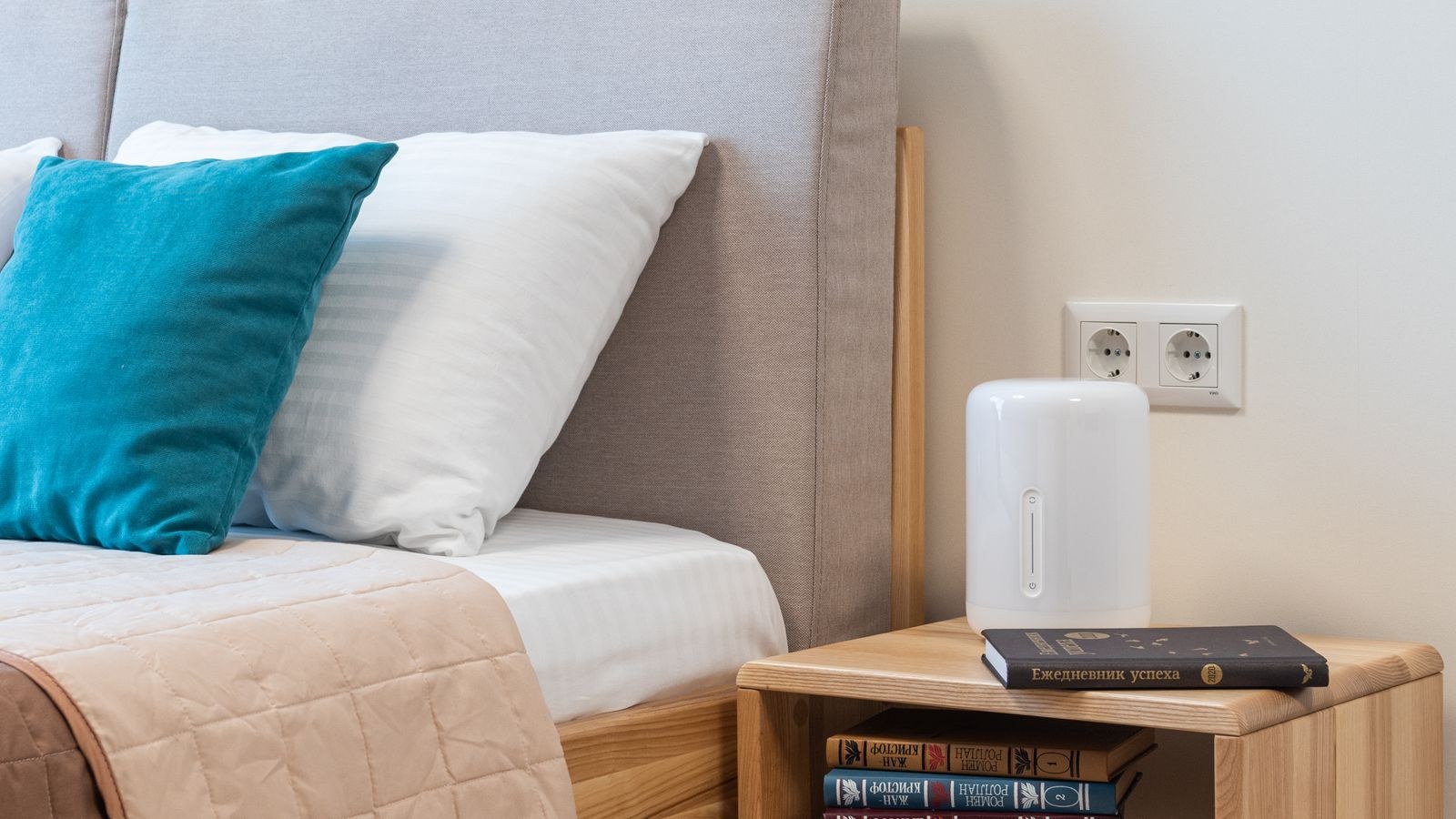 Bedside table with stacked books next to a made bed in a bright bedroom