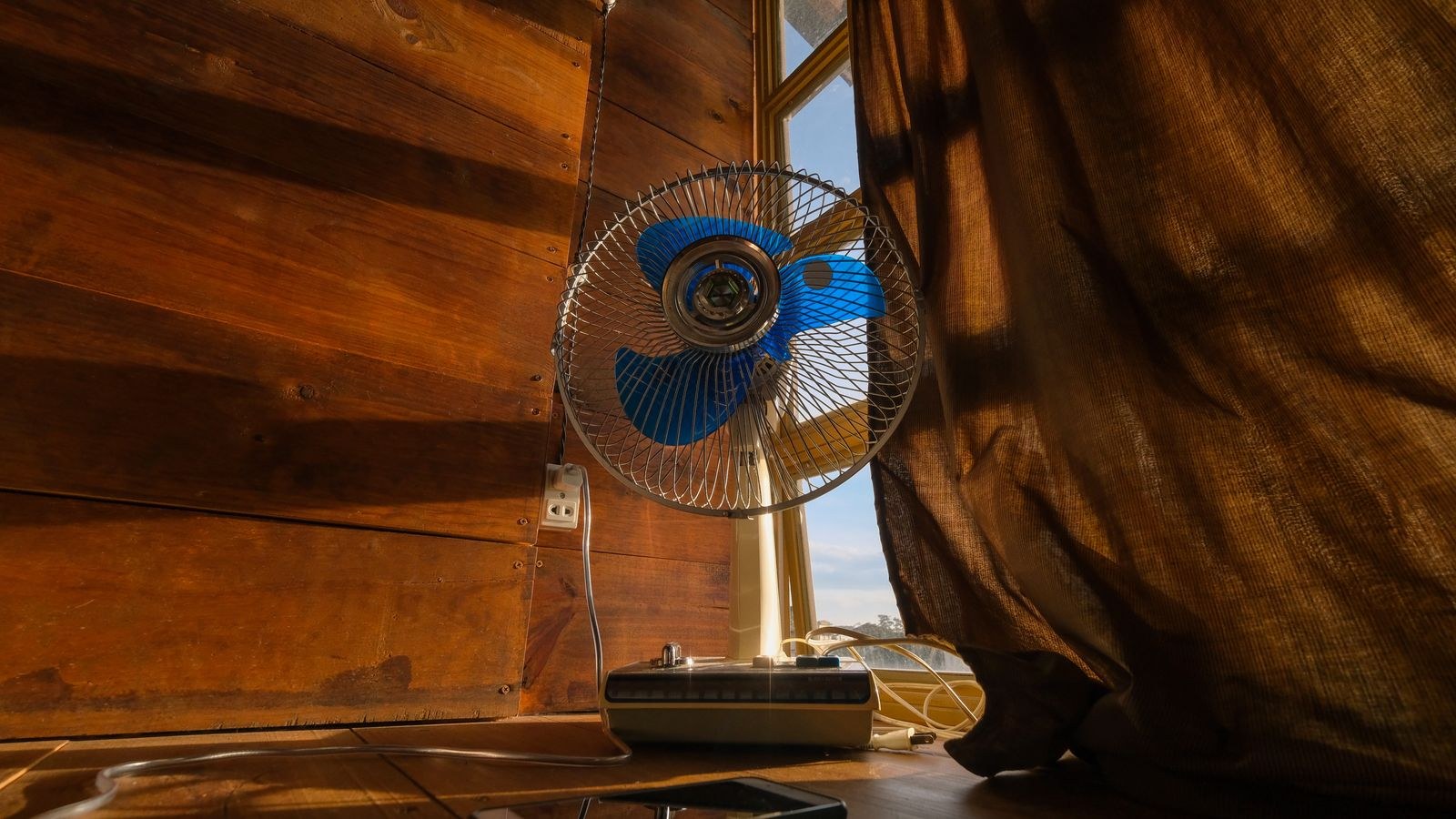 Electric fan beside a sunlit bedroom curtain with a phone charging nearby